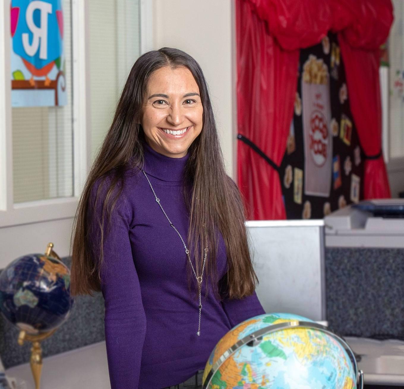 A headshot of Lauren, a teacher with long hair, wearing a long-sleeved shirt, siitting next to a globe in her classroom
