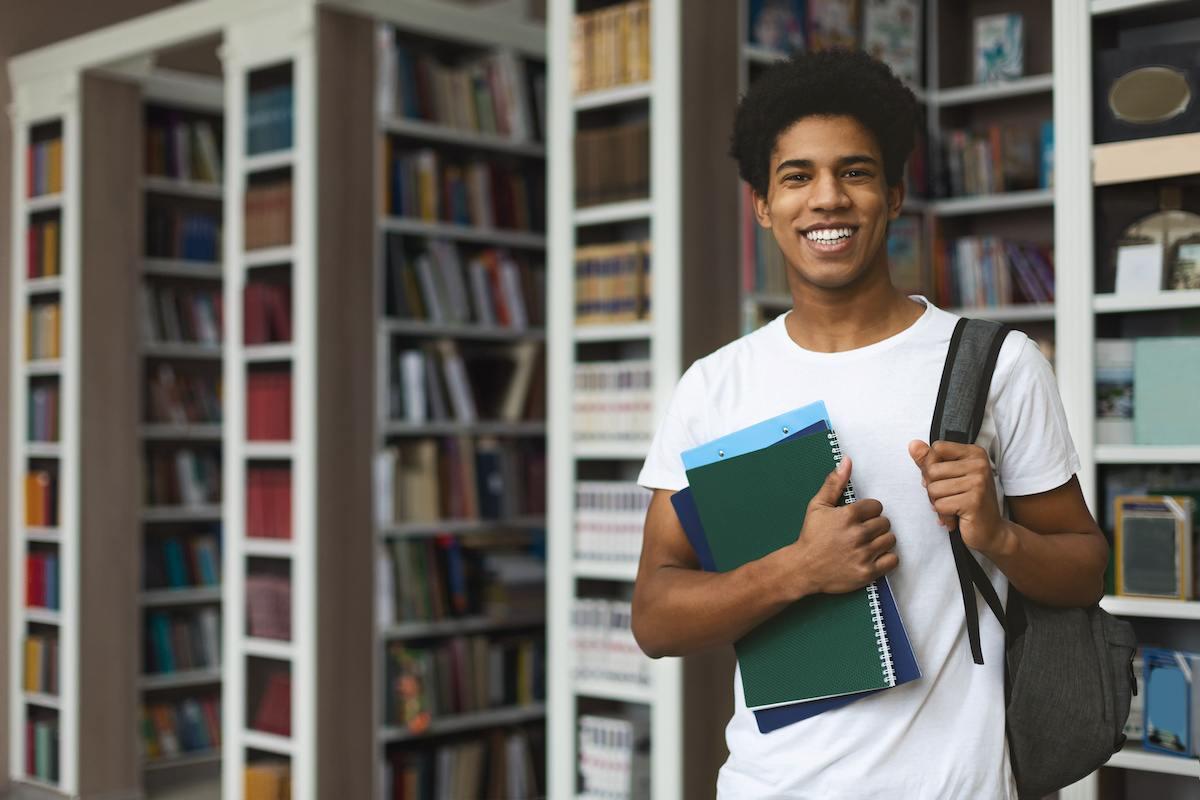 A future Connecticut teacher standing in his campus library. He’s wearing a white T-shirt and holding books in one hand and a backpack on his shoulder with the other. 